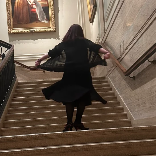 Helen in a dark dress and shawl, twirling at the foot of a grand staircase in a gallery.