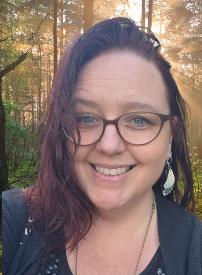 A white woman with reddish brown hair and brown eyeglasses smiles at the camera with a woodland scene at her back and the sun shining from behind.
