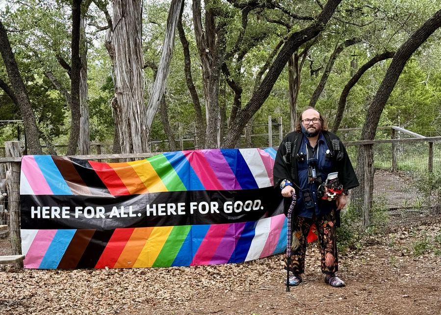Ryan standing by a Pride banner reading Here For All Here For Good.